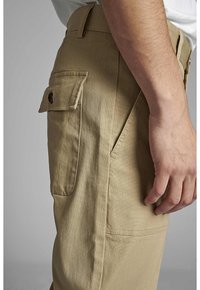 Khaki cargo kalhoty s hladkou texturou, nabízející boční kapsu se zapínáním na knoflík a šikmé kapsy na ruce. Viditelné švy.