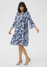 Modro-bíl dress s květinovým vzorem, zvonovými rukávy, výstřihem do V a vrstvenou sukní. Nosí se s béžovými podpatky.