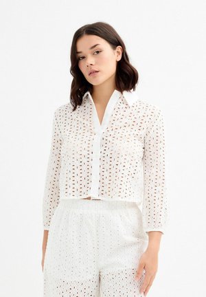 Camisa corta blanca hecha de un tejido transparente con un diseño perforado, mangas largas y un cierre frontal abotonado con un collar clásico.