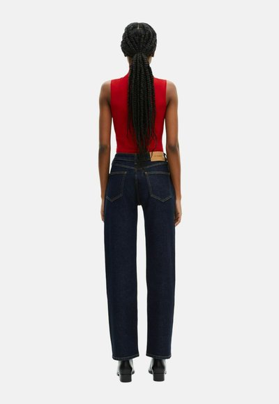 Top sin mangas rojo combinado con jeans de tiro alto y pernera ancha en azul oscuro. Los jeans cuentan con bolsillos clásicos y detalles de costura. Botas de tacón negras visibles.