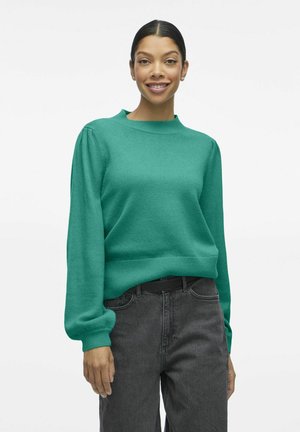 Grön stickad tröja med rund halsringning, avslappnad passform och långa ärmar med puffdetaljer. Kombinerad med högmidjade mörkgrå jeans.