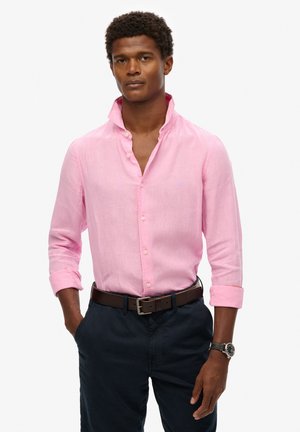 Camisa de lino rosa claro con cuello extendido, botonadura y mangas arremangadas, combinada con pantalones azul marino y un cinturón marrón.