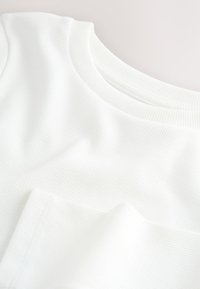 Tissu blanc côtelé avec une texture lisse, présentant un col rond et une tombée décontractée, mettant en valeur des lignes subtiles dans le matériau.