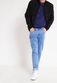 Mies, joka käyttää mustaa ruudullista takkia, sinistä paitaa, vaaleansiniset farkut ja valkoisia lenkkareita, seisoo valkoista taustaa vasten kädet taskuissa.