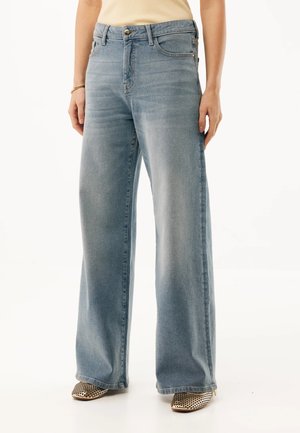 Vrouw draagt lichtblauwe jeans met wijde pijpen en voorzakken, en beige schoenen met patroon, staand tegen een effen achtergrond.