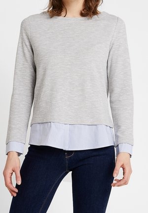 Kvinde iført en lysegrå langærmet trøje over en blå- og hvidstribet skjorte med mørkeblå jeans, stående foran en hvid baggrund.