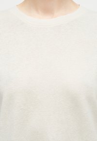 Gros plan sur une personne portant un pull à col rond uni de couleur beige clair, montrant le cou et la partie supérieure de la poitrine.