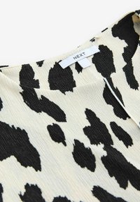 Tissu à motifs animaliers en noir et blanc, présentant une surface texturée avec un léger éclat, coupé dans un design à encolure dégagée. Étiquette de marque visible.