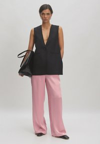 Schwarzer ärmelloser Blazer mit tiefem V-Ausschnitt, einem einzigen Knopf, kombiniert mit hellrosa weiten Hosen und einer schwarzen Handtasche. Glatte Texturen.