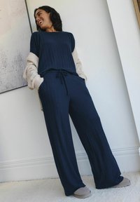 Granatowy, prążkowany komplet loungewear z sznurkiem w talii; połączony z kremowym, puszystym kardiganem z długim rękawem i szarymi kapciami na teksturowanym dywanie.