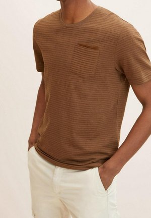 Brun kortærmet ribstrikket t-shirt med brystlomme, båret med offwhite bukser, hænder i lommer, overkrop synlig fra hals til hofter.