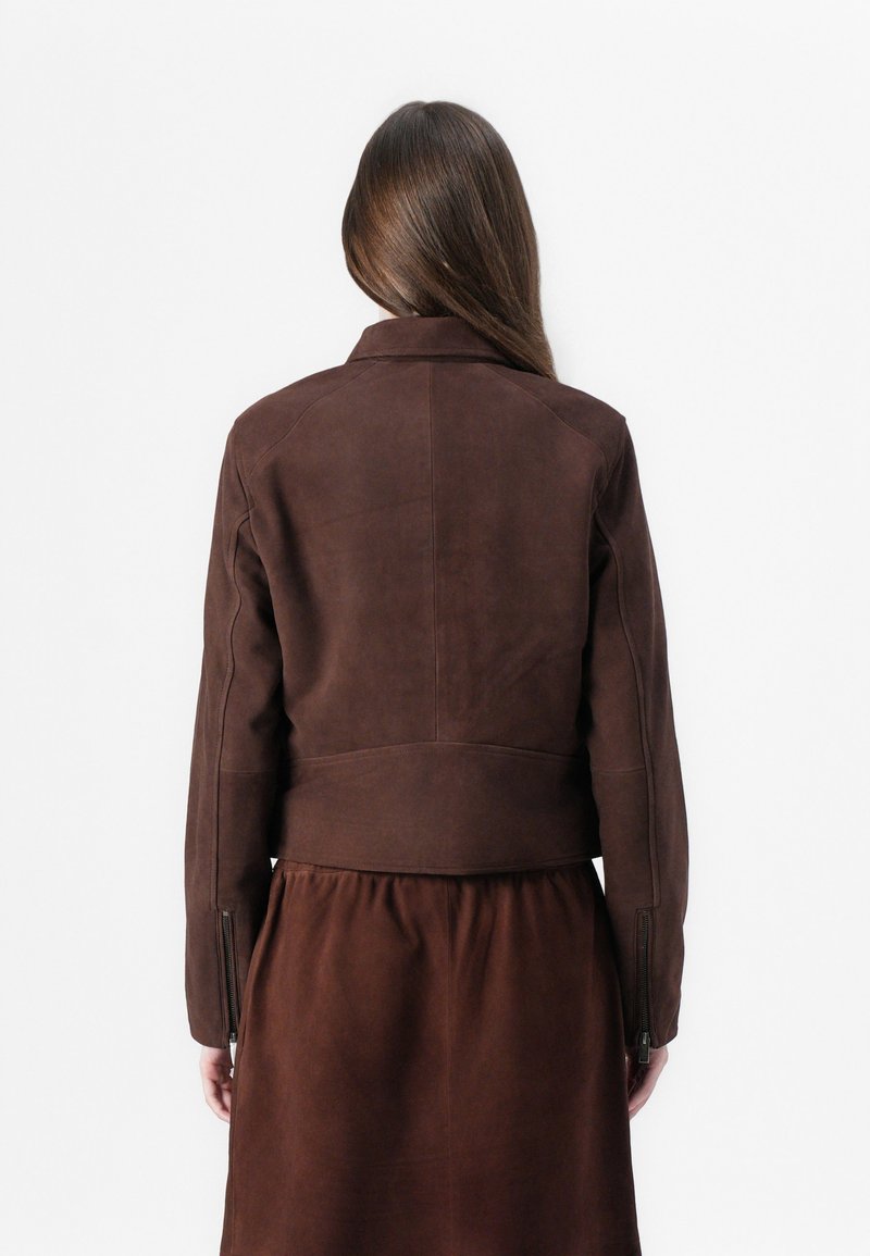 Brązowa zamszowa kurtka o krótkim kroju z kołnierzem i zapięciem na suwak, charakteryzująca się gładką fakturą i eleganckim designem, noszona na dopasowanej brązowej sukience.