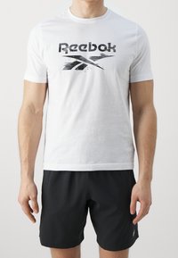 Bílý bavlněný tričko s černým logem Reebok v opotřebovaném stylu, doplněné černými sportovními šortkami.
