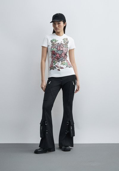 Νεαρή γυναίκα που φοράει άσπρο γραφικό μπλουζάκι με σχέδιο σφαίρας, μαύρο παντελόνι καρό με τσέπες, μαύρα παπούτσια και μαύρο καπέλο μπέιζμπολ.