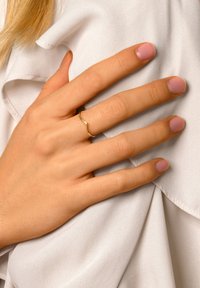 Anillo de color dorado con un diseño ondulado colocado sobre una mano. Las uñas están bien cuidadas con un esmalte rosa claro. La mano descansa sobre una tela suave y blanca.
