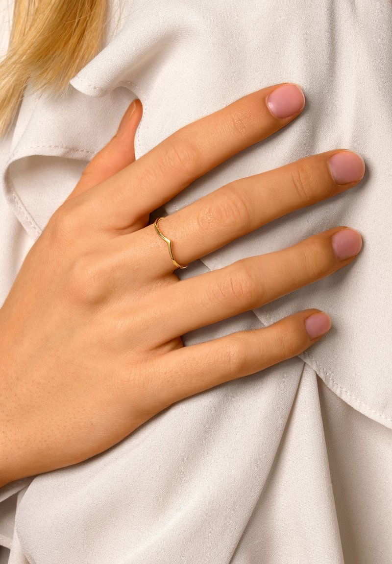 Anillo de color dorado con un diseño ondulado colocado sobre una mano. Las uñas están bien cuidadas con un esmalte rosa claro. La mano descansa sobre una tela suave y blanca.