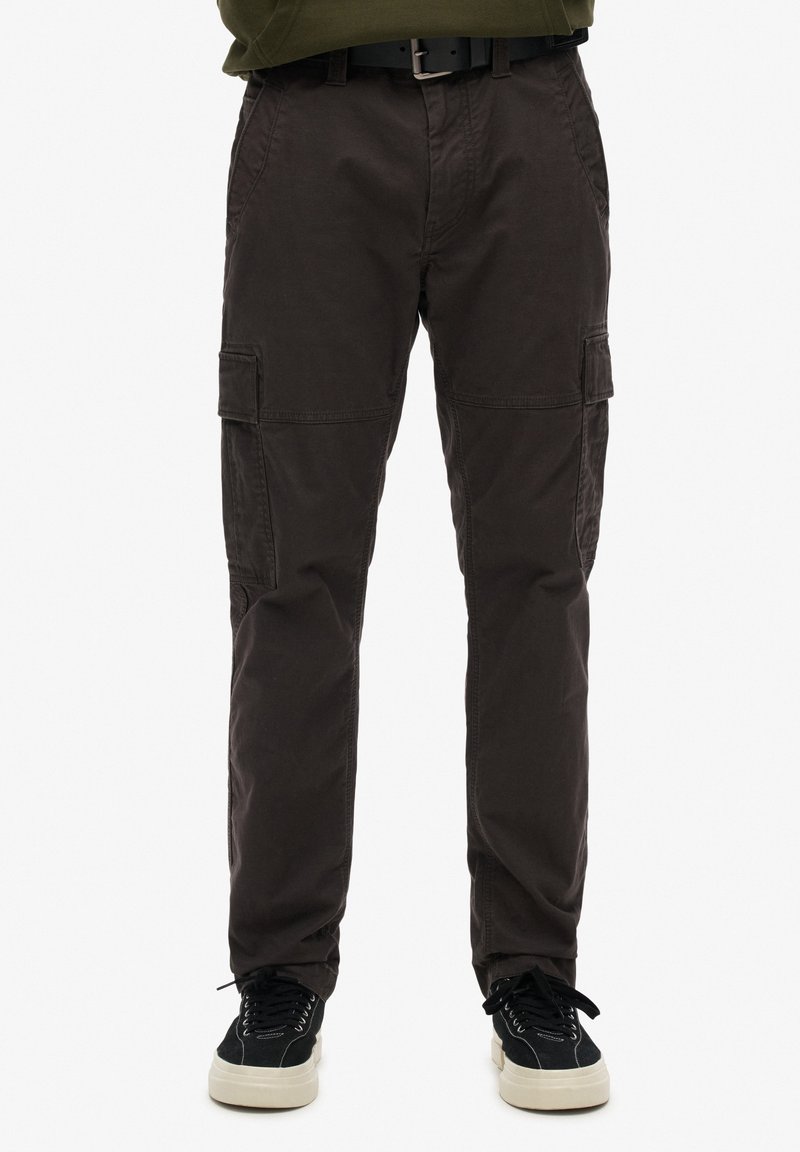 Homme portant un pantalon cargo marron foncé avec des poches à rabat et des baskets noires aux semelles blanches sur un fond uni.