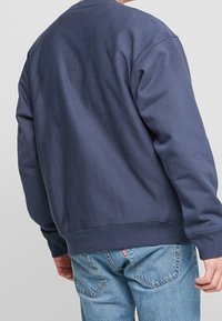 Man i en enkel marinblå sweatshirt och ljusblå denimjeans, sedd bakifrån mot en enkel grå bakgrund.