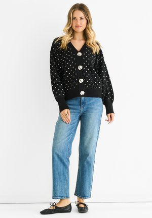 Black knitted cardigan with white polka dots, featuring a deep V-neck and five large decorative buttons. Paired with straight-leg blue jeans.