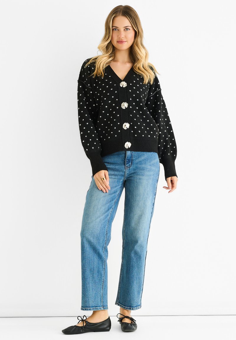 Black knitted cardigan with white polka dots, featuring a deep V-neck and five large decorative buttons. Paired with straight-leg blue jeans.