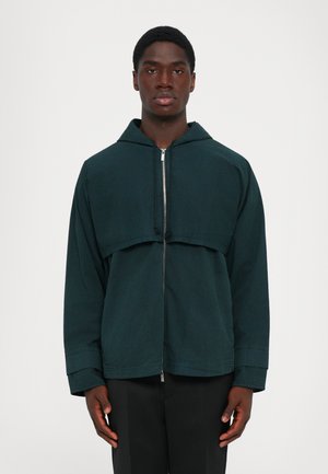 Młody mężczyzna w ciemnoniebieskiej kurtce z zamkiem i kapturze oraz czarnych spodniach, stojący na tle jednolitego białego tła.