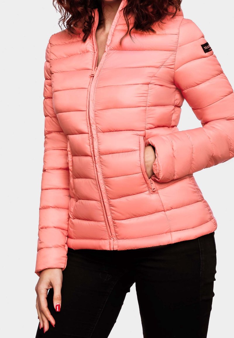 Femme portant une veste matelassée corail avec des poches zippées et un pantalon noir, une main dans la poche et l'autre reposant à côté d'elle.