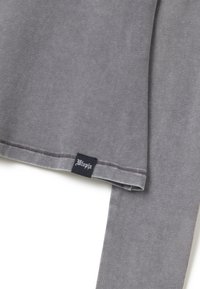 Top lungo in cotone grigio con maniche lunghe, scollo rotondo, cuciture in evidenza e un'etichetta nera con scritte bianche sul orlo.