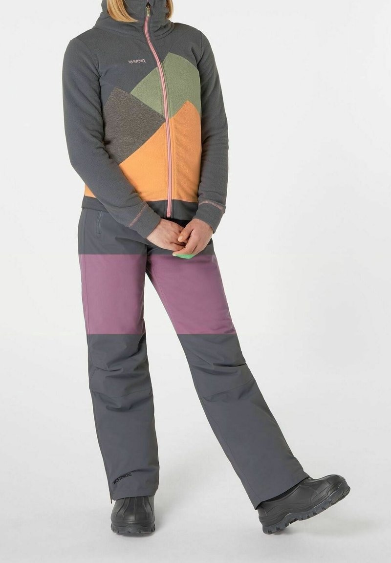 Personne portant une veste zippée colorée et un pantalon de plein air foncé avec des bottes noires, debout avec une jambe croisée sur l'autre.