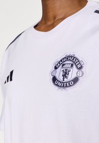 Bílé bavlněné tričko s černobílým logem Manchester United na hrudi a černými proužky na ramenou.