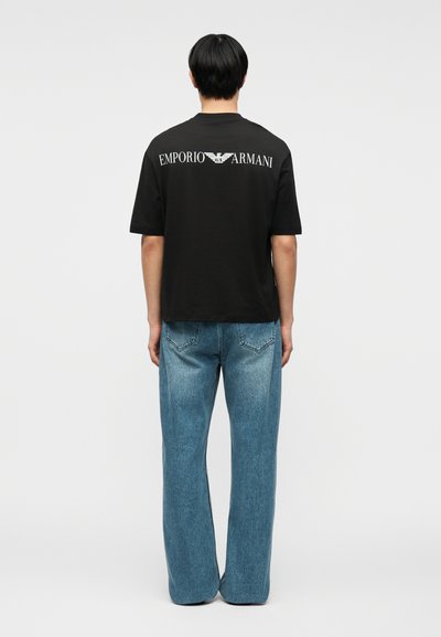 Άνδρας που φορά μαύρο μπλουζάκι Emporio Armani και μπλε τζιν, στέκεται με την πλάτη προς το μέρος μας μπροστά σε απλό ανοιχτό γκρι φόντο.
