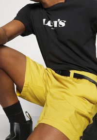 Černé tričko s logem "Levi's", spárované se žlutými šortkami s kapsami a černým opaskem. Viditelné černé ponožky a lesklé černé kotníkové boty.