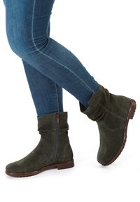 Persona che indossa stivaletti in camoscio verde scuro con cerniere laterali e jeans skinny blu, incrociando una gamba sull'altra.