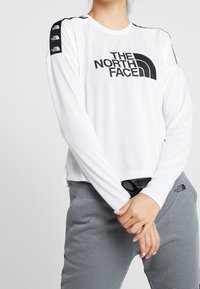 Person som bär en vit långärmad tröja från The North Face och gråa sweatpants från The North Face, med händerna knäppta framför sig.