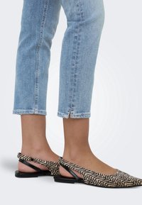 Personne portant un jean court bleu clair et des chaussures plates slingback beiges et noires à motif zigzag sur fond uni.