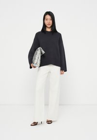 Dunkelgrauer, übergroßer Strickpullover, weit geschnittene weiße Hose, hält eine metallische Clutch im Schlangenprint und trägt schwarze, schulterfreie Sandalen mit Absätzen.