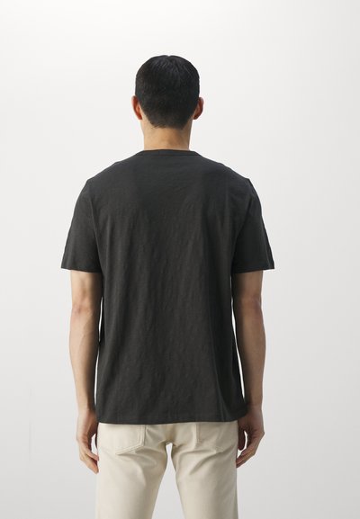 Black crew-neck T-shirt made from a textured fabric, featuring short sleeves and a relaxed fit, worn with light beige trousers.