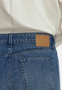 Blå denimjeans med en tan läderetikett märkta "FALCONERI" på den bakre midjan; synlig söm och en höger bakficka.