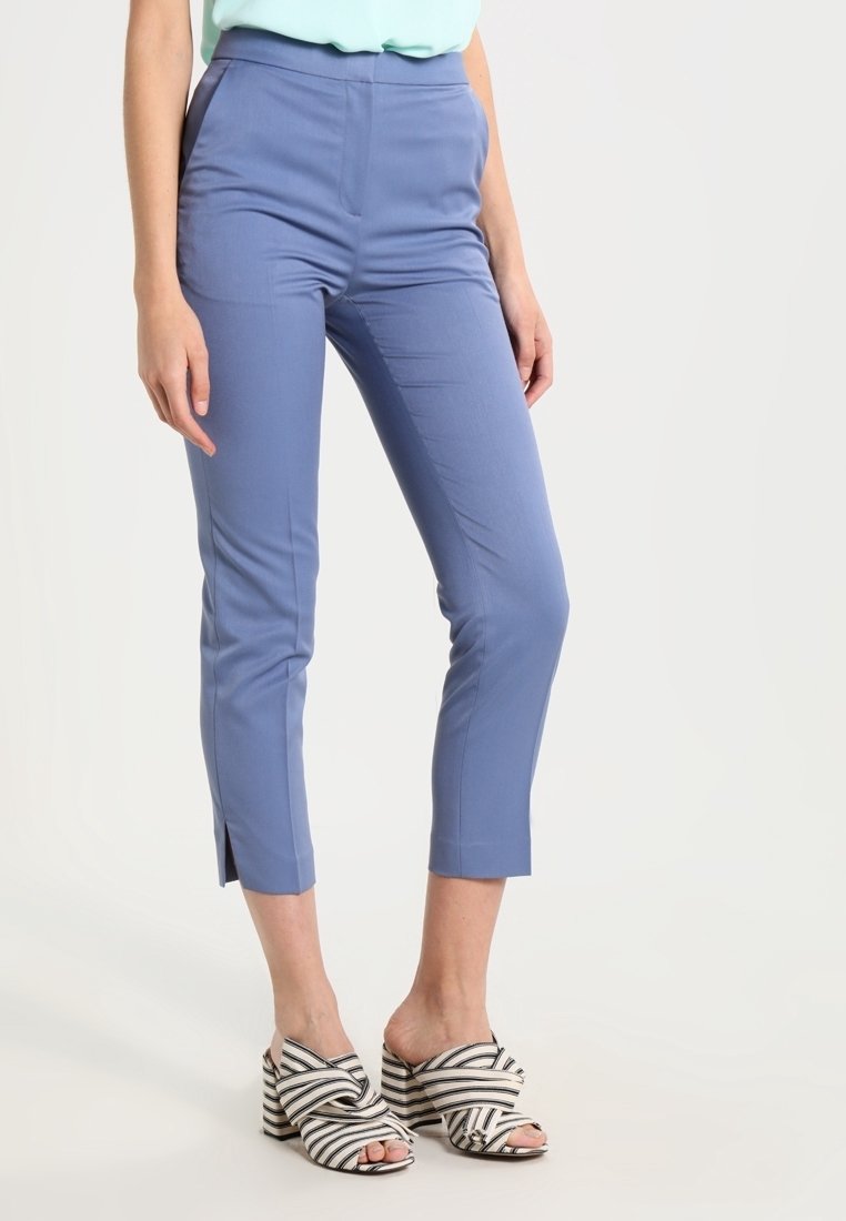 Femme portant un pantalon court slim bleu clair et des sandales à talons rayées noir et blanc, debout devant un fond uni.