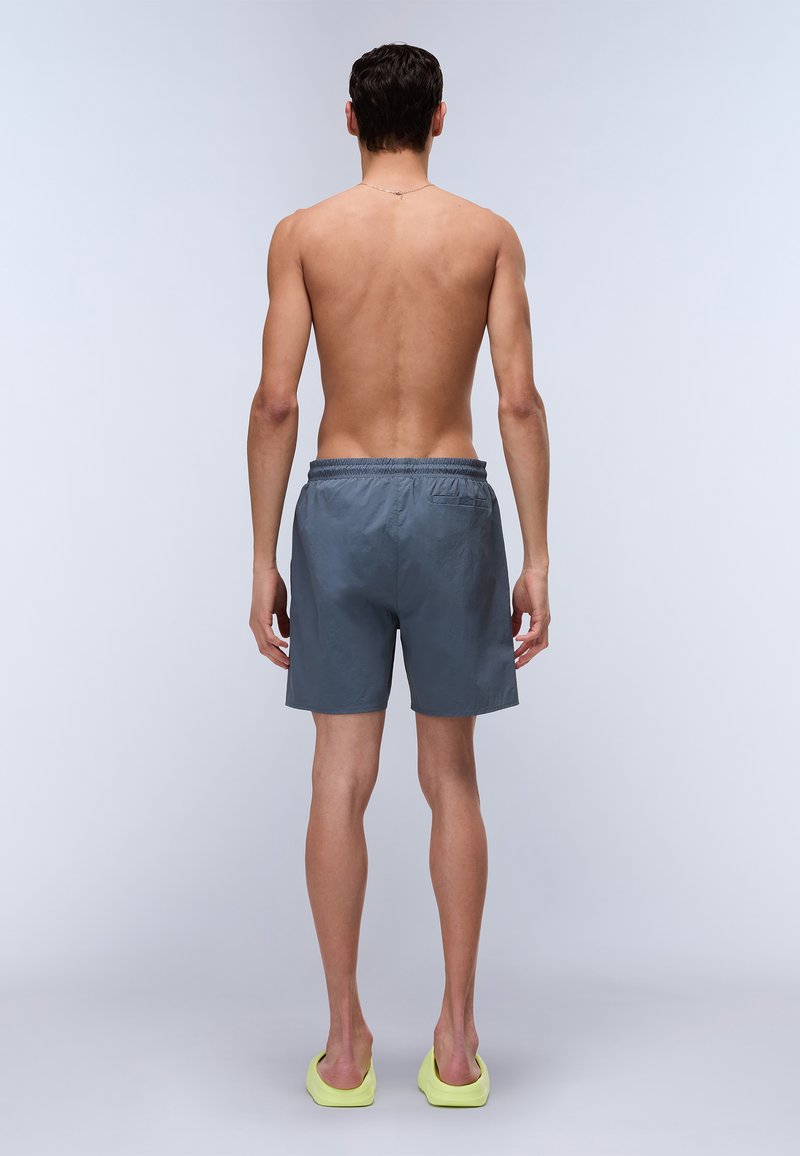 Shorts de bain gris en matériau léger, avec une taille élastique, une coupe courte et une poche arrière. Portés avec des sandales à glisser jaunes.