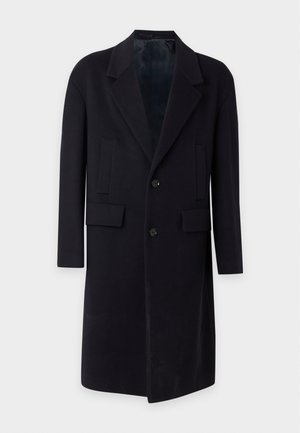 Homme grand portant un long manteau noir par-dessus un polo gris, pantalon noir et chaussures noires, debout devant un fond clair uni.