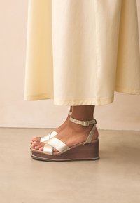 Ék talpú szandál texturált arany felsőrészsel, bokapánttal és fa talppal. A talp sötét, ami kontrasztot alkot a felső rész színével.