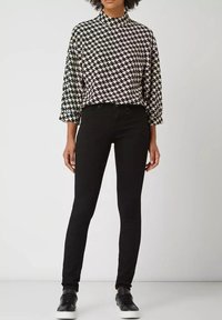 Sort houndstooth-mønsteret top med høj krave og trekvartærmer, parret med tætsiddende sorte jeans og sorte sneakers.