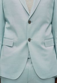 Ανοιχτό μπλε σακάκι κοστούμι με δύο κουμπιά κλειστά, τσέπες με καπάκια και ταιριαστό παντελόνι.