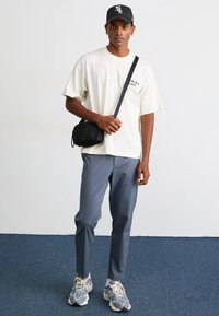 Cream cotton t-shirt with printed text, gray tapered pants, black crossbody bag, and gray sneakers with purple accents. Plain background.