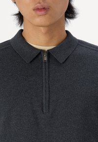 Mörkgrå zip-up polo skjorta med krage, ribbad textur och en kort dragkedja framtill. Under finns ett ljusbeiget lager.