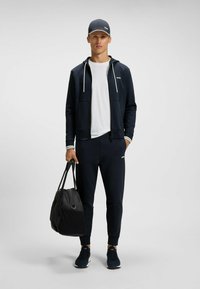Felpa a zip blu marina con lacci bianchi, abbinata a una maglietta bianca e pantaloni jogger blu marina coordinati. Mostrata una borsa da viaggio nera e scarpe da ginnastica scure.