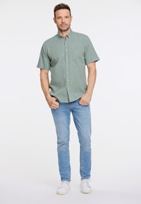 Camicia verde a maniche corte con bottoni e taschino sul petto, abbinata a jeans blu chiaro e sneaker bianche, in piedi davanti a uno sfondo neutro.