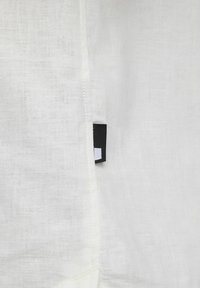 Balta lino medžiaga su tekstūru, su juodu etikete, ant kurios siuvinėtas baltas logotipas. Minimalistinis dizainas ir lengvas.