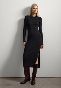 Schwarzes, langärmliges, geripptes Kleid mit einem seitlichen Schlitz, aus einem glitzernden Material. Kombiniert mit hohen, glänzenden burgunderfarbenen Stiefeln.