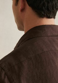 Camisa de lino marrón con una textura suave, cuello rectangular y hombros estructurados, vista desde la parte posterior, que resalta los detalles de la costura.